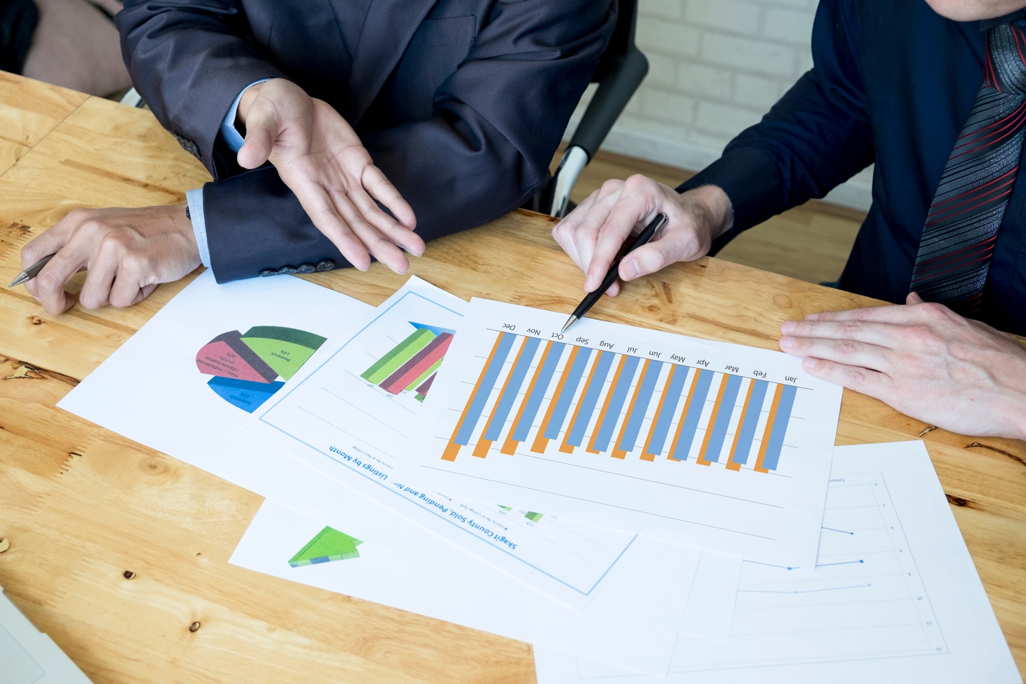 Two business professionals in suits analyze financial charts and graphs spread across a wooden table, discussing trends and data insights.